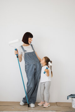 A mother and her young daughter stand side-by-side, holding paint rollers and looking at each other with smiles as they prepare to paint a room