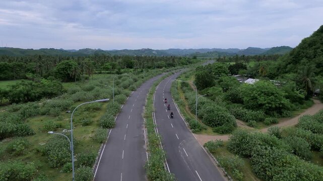 Aerial Winding Road Through Scrub And Bushes, Empty Lanes Curve Toward Distant Hills, Lampposts And Signage Align Roadside, Overgrown Verges And Quiet Cloudy Sky, Conservation Mapping Tone