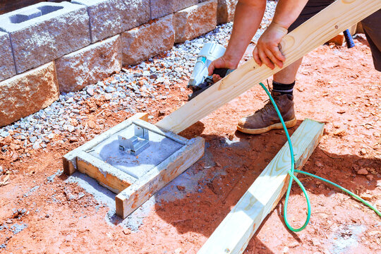 Construction worker secures wooden board to concrete base using nail gun during building project outdoors.