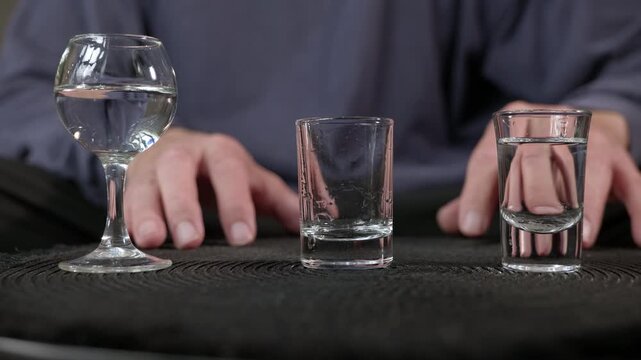 Person drinking several alcoholic shots from glasses. Man's hands picking up and drinking shots of a clear alcoholic beverage from different glasses on a black table, representing a alcoholism