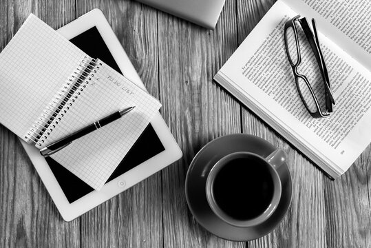 Flat lay of cozy workspace with laptop, red cup of black coffee, open book with glasses and notepad on wooden desk