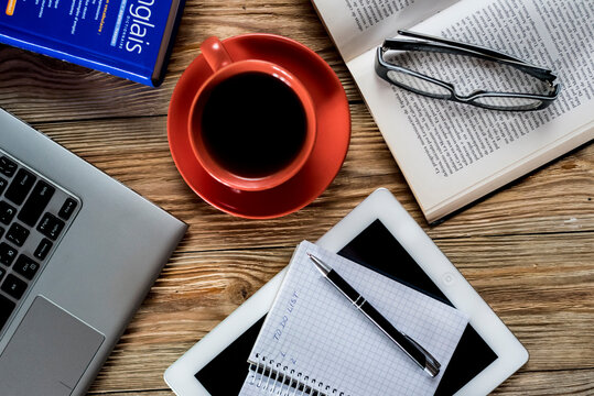 Flat lay of cozy workspace with laptop, red cup of black coffee, open book with glasses and notepad on wooden desk