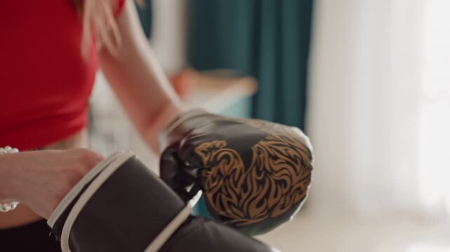 Home fitness white woman fastening gloves closeup showing velcro straps, black gloves with gold tiger motif, red crop top and bracelet, living room light, calm focused preparation for athome training