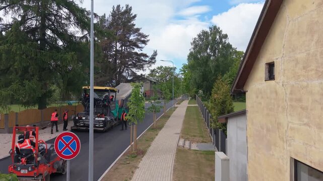 Road construction crew operates asphalt paving machinery on residential street, showcasing continuous action and teamwork in urban setting