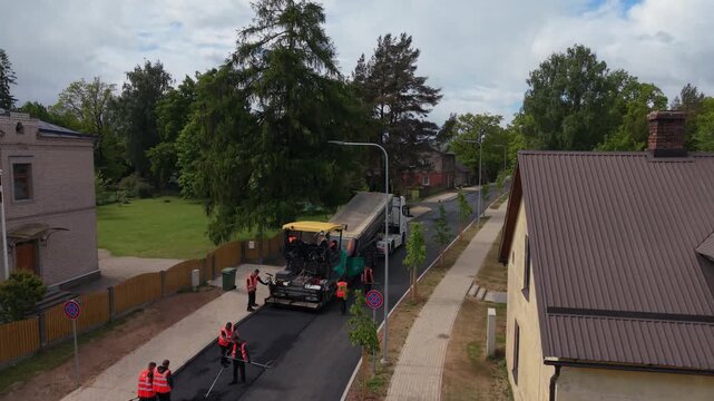 Road construction team lays asphalt on a residential street with machinery and workers in safety vests visible in the background