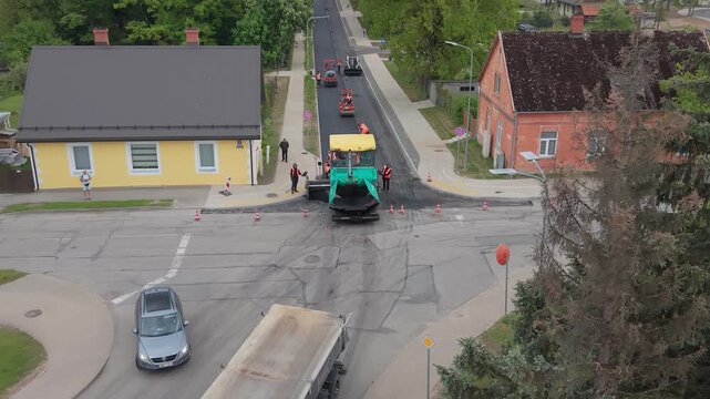 Road construction crew operates asphalt paver and roller on street intersection with traffic cones and vehicles in view