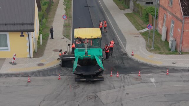 Road construction crew operates asphalt paver on street, laying fresh asphalt while traffic cones and buildings are visible nearby