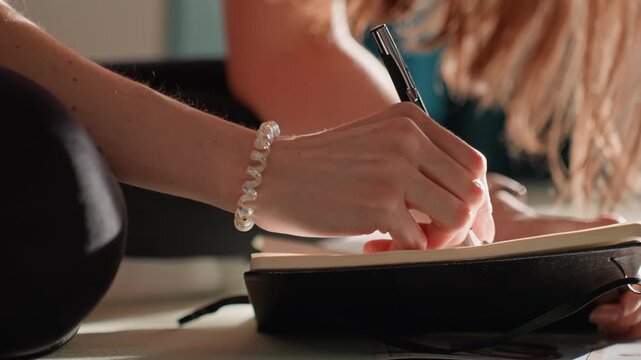 Caucasian woman writing fitness journal on mat, black notebook and pearl bracelet under warm sunlight, pen strokes marking workout plan, cozy living room floor, closeup hands crafting weekly routine