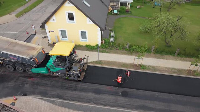 Road construction workers in safety vests operate asphalt paver and prepare fresh asphalt surface near residential house and green lawn
