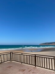 Fototapeta premium Wooden terrace overlooks wide sandy beach with people walking near Atlantic Ocean waves on sunny day in Portugal