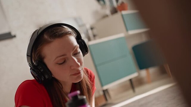 Young woman home workout session headphones, red shirt, purple water bottle visible, indoor lounge setting with soft natural light, following online trainer audio, focused breathing and gentle