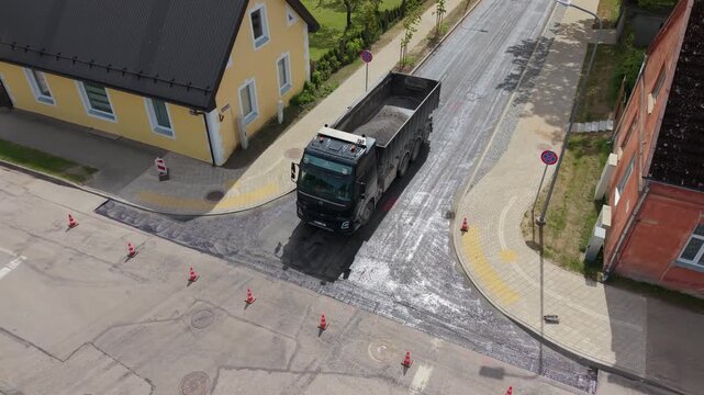 Truck transporting gravel navigates a residential street with yellow house and roadwork cones visible in the background