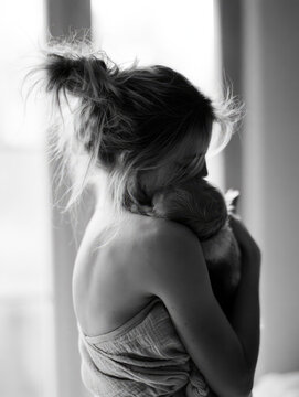 Postpartum mother hugging newborn back view messy hair bun tired posture natural light close up black and white breastfeeding top soft fabric intimate family moment