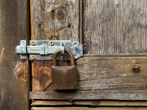 Old metal padlock attached to a wooden door latch on a rustic farm building. Detailed view of traditional locking hardware, weathered timber and countryside architecture, symbolizing security