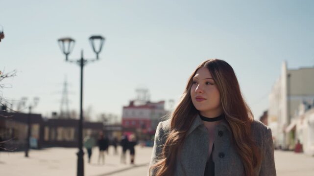 Pretty caucasian woman facing camera in plaza, fashion model vibe, gray coat, gentle gaze, lamp posts in background, soft sunlight, candid influencer movement, thoughtful expression