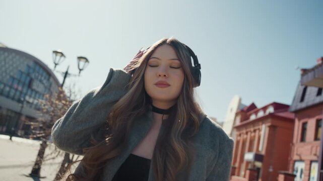 Sunlit city street portrait of woman in coat and beanie, headphones on, looking up at glass market and redbrick buildings peaceful expression, cinematic wideangle, soft cool tones, gentle breeze