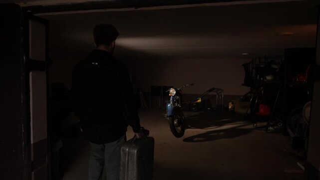 Man holding a metal fuel canister, illuminating a vintage motorcycle with a flashlight beam in a dark garage, preparing for a potential journey or maintenance