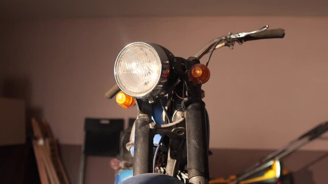 Vintage motorcycle headlight, handlebars and front fork with chrome patina and turn signal, close up view of a classic bike mid restoration in a home garage workshop setting