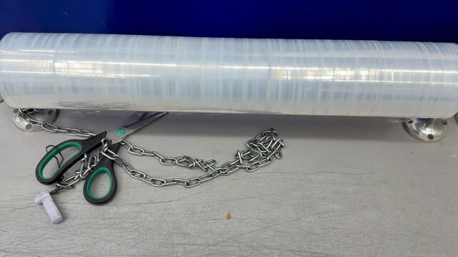 Clear stretch film roll on counter, packaging station with scissors, tape dispenser and metal chain, textured tabletop background