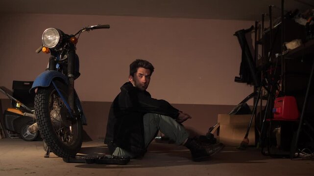 Mechanic man sitting on a garage floor next to an old motorcycle, looking at the camera, thinking about problems or solutions while working on vehicle repair