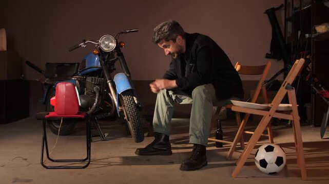 Man sitting in a dimly lit garage, intently watching a soccer game on an old analog television while a vintage motorcycle and a soccer ball are nearby, evoking a sense of nostalgia