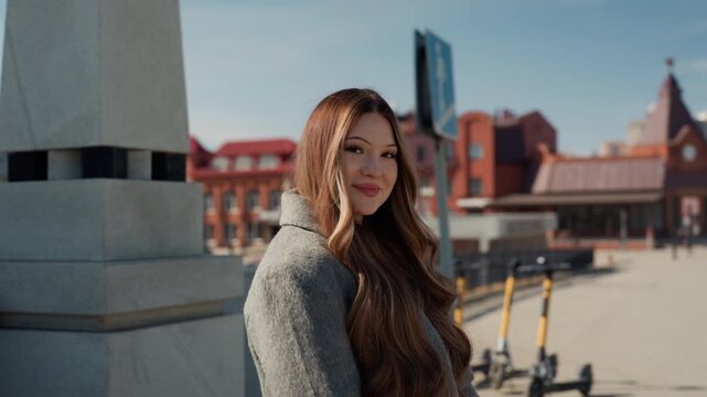 Pretty asian traveler at station smiling near plaza architecture, relaxed pose in wool coat, long hair framing face, redbrick buildings and glass station in background, sunny day mood with friendly