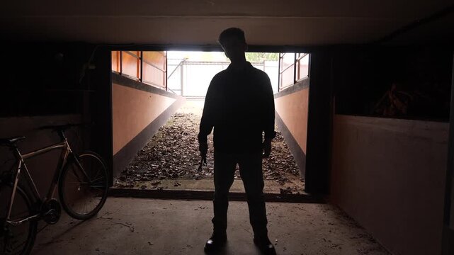 Man holding a wrench standing silhouetted against a bright background, creating a mysterious and atmospheric scene in a dimly lit, abandoned looking garage with a bicycle
