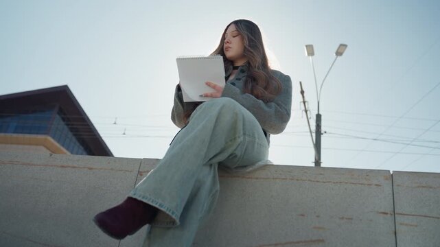 Pretty asian illustrator sketching on concrete wall at street edge, lamp posts and building behind, relaxed pose in jeans and coat, clear sky and soft daylight give calm creative atmosphere