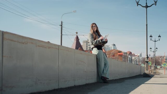 Pretty white woman standing by wall writing journal city notes, traveler journaling through streets, observer noticing rooftops and lampposts, casual denim, sunlight shadow play, reflective mood