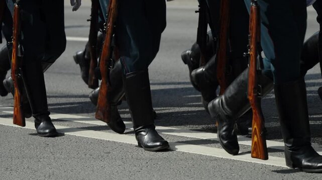 Close-up view of uniformed soldiers marching in formation along an asphalt street, black boots stepping in unison with wooden-stock rifles held at their sides during a parade.
