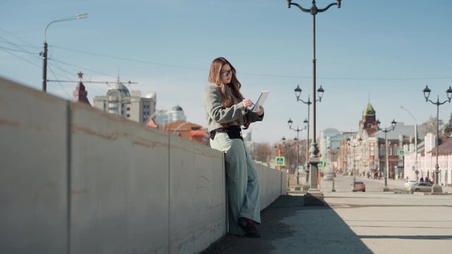 Pretty white woman leaning on wall checking tablet, designer planning layouts, glasses on, sketchbook nearby, focused expression, lamp posts and city spires in background, soft daylight