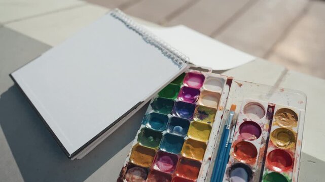 Detailoriented workspace with shimmering art supplies. Calm and cozy creative scene with watercolor tools. Morning light highlights textured paper and glossy paint pans in tranquil studio setting