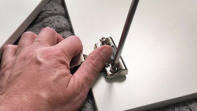 Close-up of a hand tightening screws on a concealed furniture hinge with a screwdriver, demonstrating cabinet door assembly, installation, adjustment, and repair work