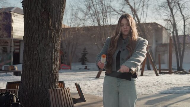 Pretty woman removing lint from coat sleeve, outdoor winter park scene with sunlit snow, curly hair framing face, denim pants and wool coat, methodical rolling motion with lint roller, student