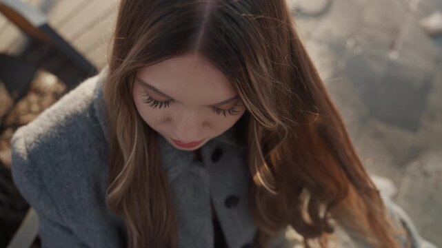 Overhead winter portrait of pretty caucasian, young woman in gray coat on park bench, long wavy hair dusted with snowflakes, hand brushing hair, soft golden light, introspective mood, cinematic