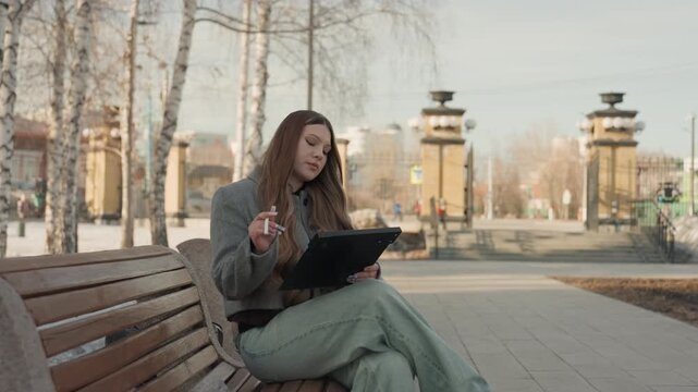 Pretty white woman on bench sketching on tablet and planning designs scrolling reference images, drawing concept lines, adjusting layers, reviewing portfolio, saving files. Long brown hair, gray coat,