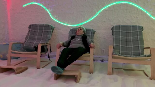 Elderly woman relaxing in salt cave halotherapy chamber, wearing cap and reclining on wooden chair with footstool, soft neon wave