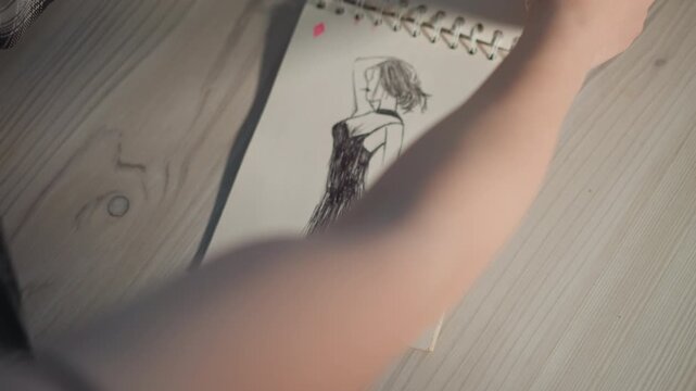 Designer finalizing dress sketch on notebook, refined ink silhouette and contour lines, arm reaching to turn page, closeup on wooden tabletop, calm ambient lighting, fashion refinement mood.