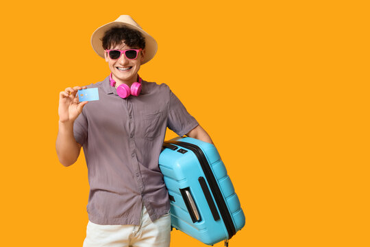 Young man in sunglasses with credit card and suitcase on yellow background