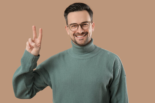 Handsome young man in eyeglasses showing victory gesture on brown background