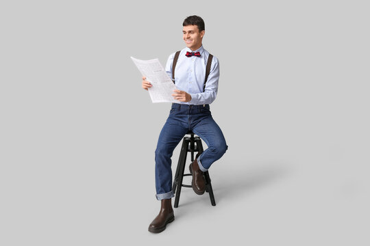 Happy young man with bow tie sitting on chair and reading newspaper on white background