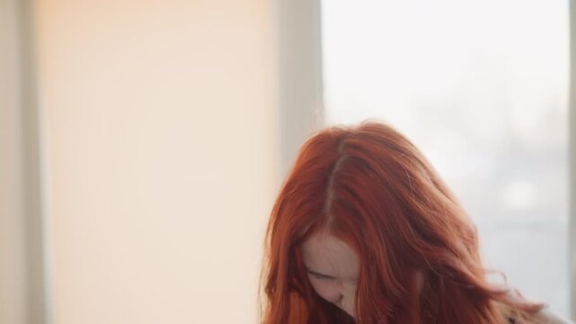 Expressive woman enjoying music. Woman with fiery red hair dancing passionately. Redheaded woman passionately headbanging near sunlit glass with energetic movement and expressive emotion