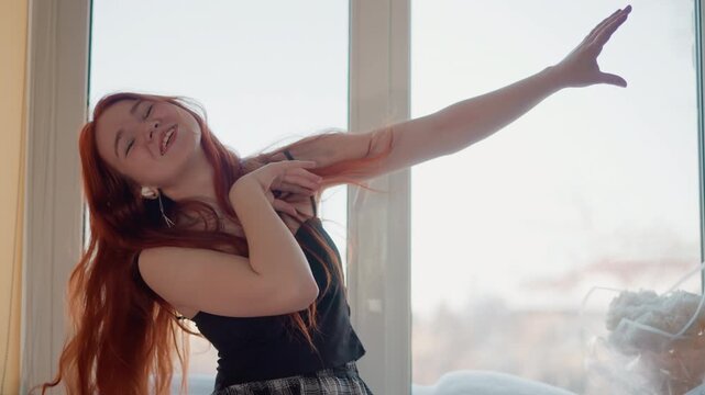 Redheaded lady moves brightly near window. Playful redhaired girl grooves cheerfully in daylight by window. Energetic woman with crimson hair dances lively outside home under bright sunlight