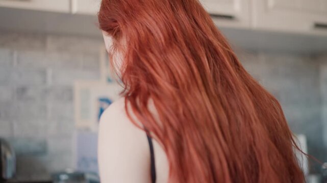 Backlit kitchen closeup caucasian woman turning toward sink, long red locks cascading over shoulders, brick backsplash and magnets blurred, peaceful postritual mood and tactile hair texture