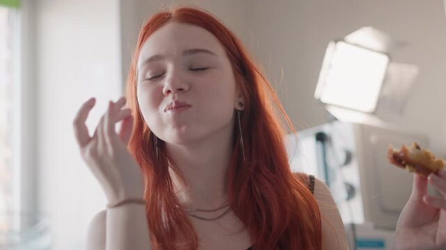 Lady relishes delight. Joyful woman with fiery hair savors dessert beneath gentle illumination. Woman with striking scarlet hair delightfully indulges in pastry under soft glowing light