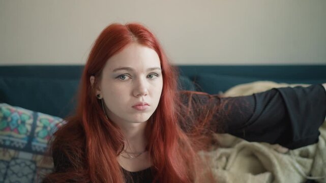 Redhead caucasian woman on sofa gazing into distance with handheld mirror and lipstick, soft blanket behind and patterned cushion frames show contemplative friend weighing choices, private oracle