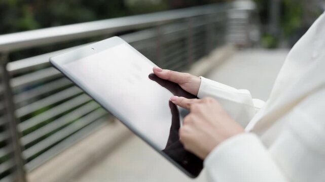 A woman in a white coat uses a tablet while standing on a modern bridge in an urban environment, showcasing a blend of technology and city life.