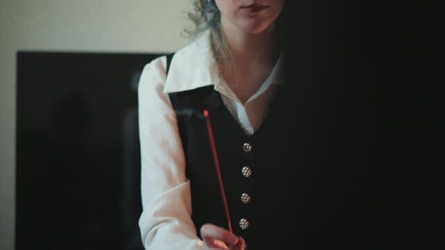 Solemn woman holding burning incense stick before dark backdrop, serious expression, formal vest, composed posture, ceremonial atmosphere, low lighting, concentrated energy, prophetic mood,