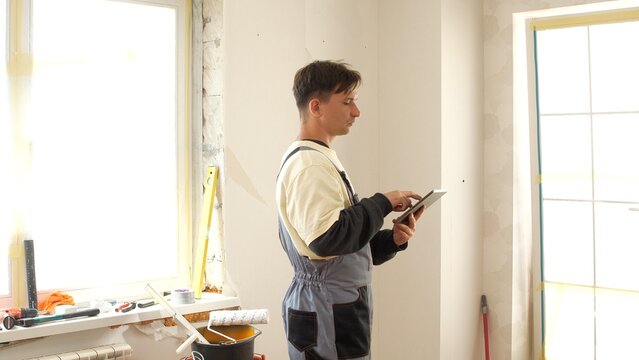 Construction worker in overalls holding and tapping digital tablet, planning and managing renovation work in a bright room with tools on the windowsill