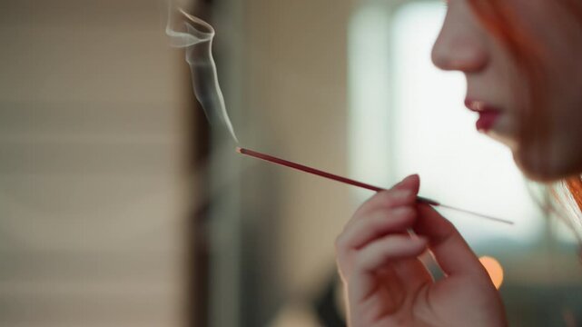 Serene fragrant ritual. Calming smoke and warm illumination. Gentle woman performs soothing cleansing with fragrant smoke. Woman breathes out incense smoke beside softly glowing window light